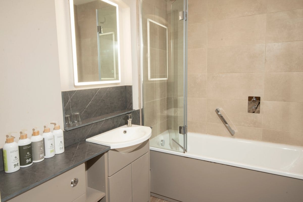 Modern bathroom with a sink, illuminated mirror, toiletries, and a bathtub with a glass screen and beige tiles.