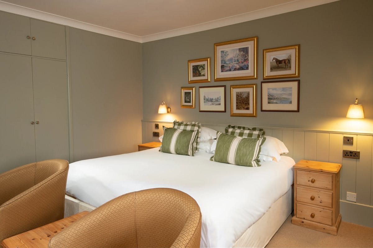 Cosy bedroom with a double bed, green pillows, wall art, and warm lighting. Two armchairs and wooden nightstands complete the decor.