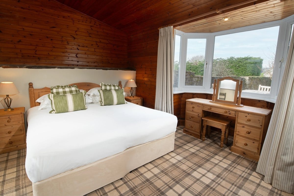 Cosy bedroom with wood panelling, a large bed, green pillows, bedside lamps, and a window-side wooden vanity with a mirror.