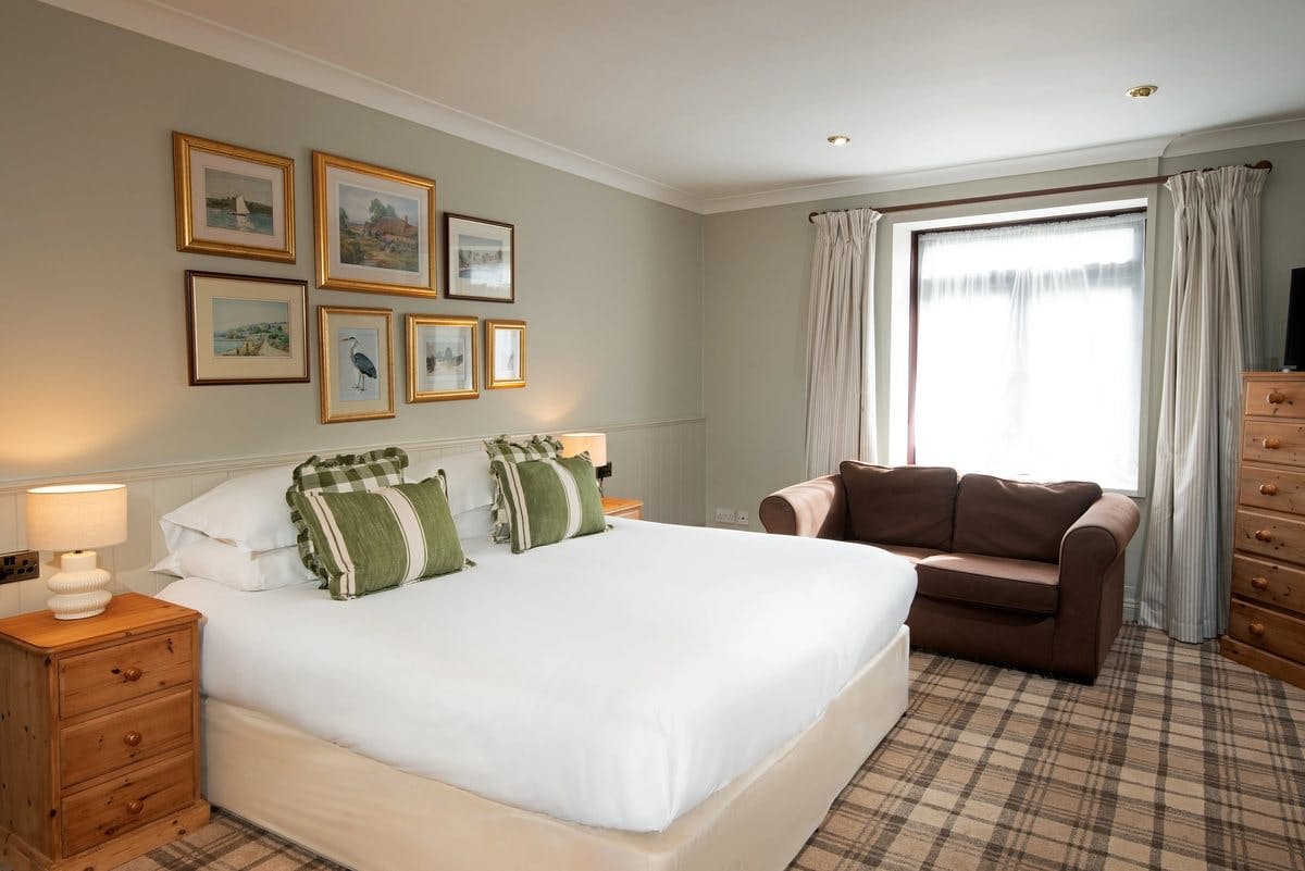 Cosy bedroom with a double bed, green pillows, brown sofa, framed art, and wooden furniture. Soft lighting and plaid carpet.