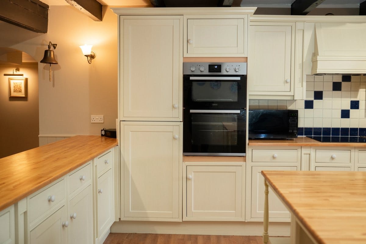 Modern kitchen with cream cabinets, wooden countertops, built-in oven, and tiled backsplash. A wall light and bell are visible.