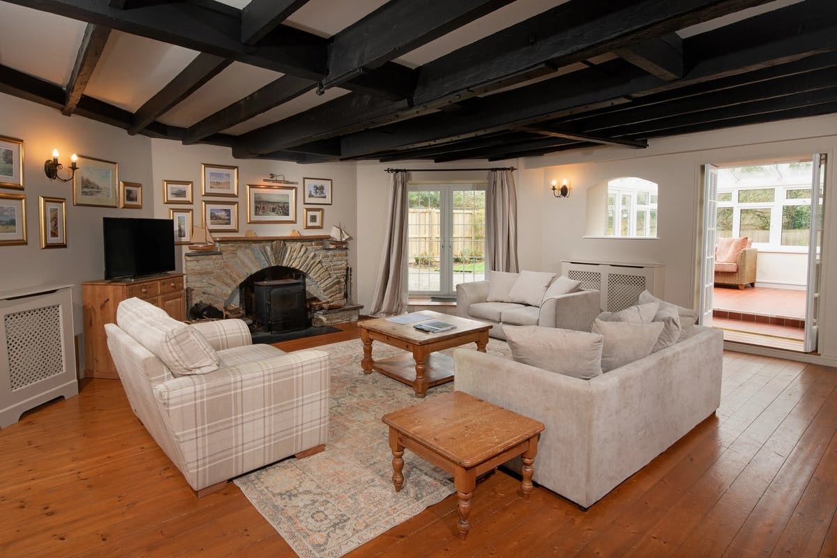 Cosy living room with sofas, a stone fireplace, wooden beams, and a TV. French doors open to a sunlit conservatory.