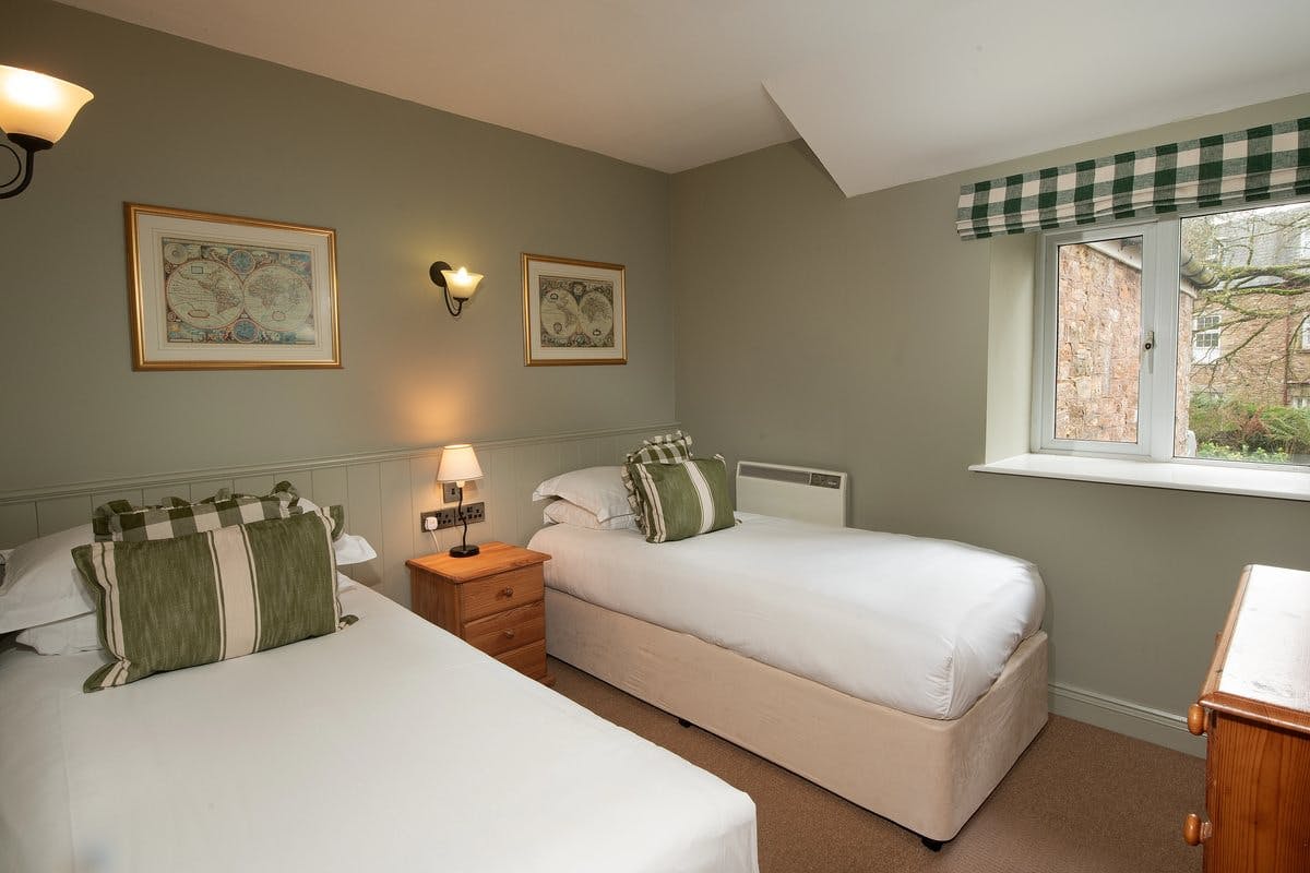 Cosy bedroom with two single beds, green pillows, a nightstand with a lamp, wall art, and a window with a checkered valance.