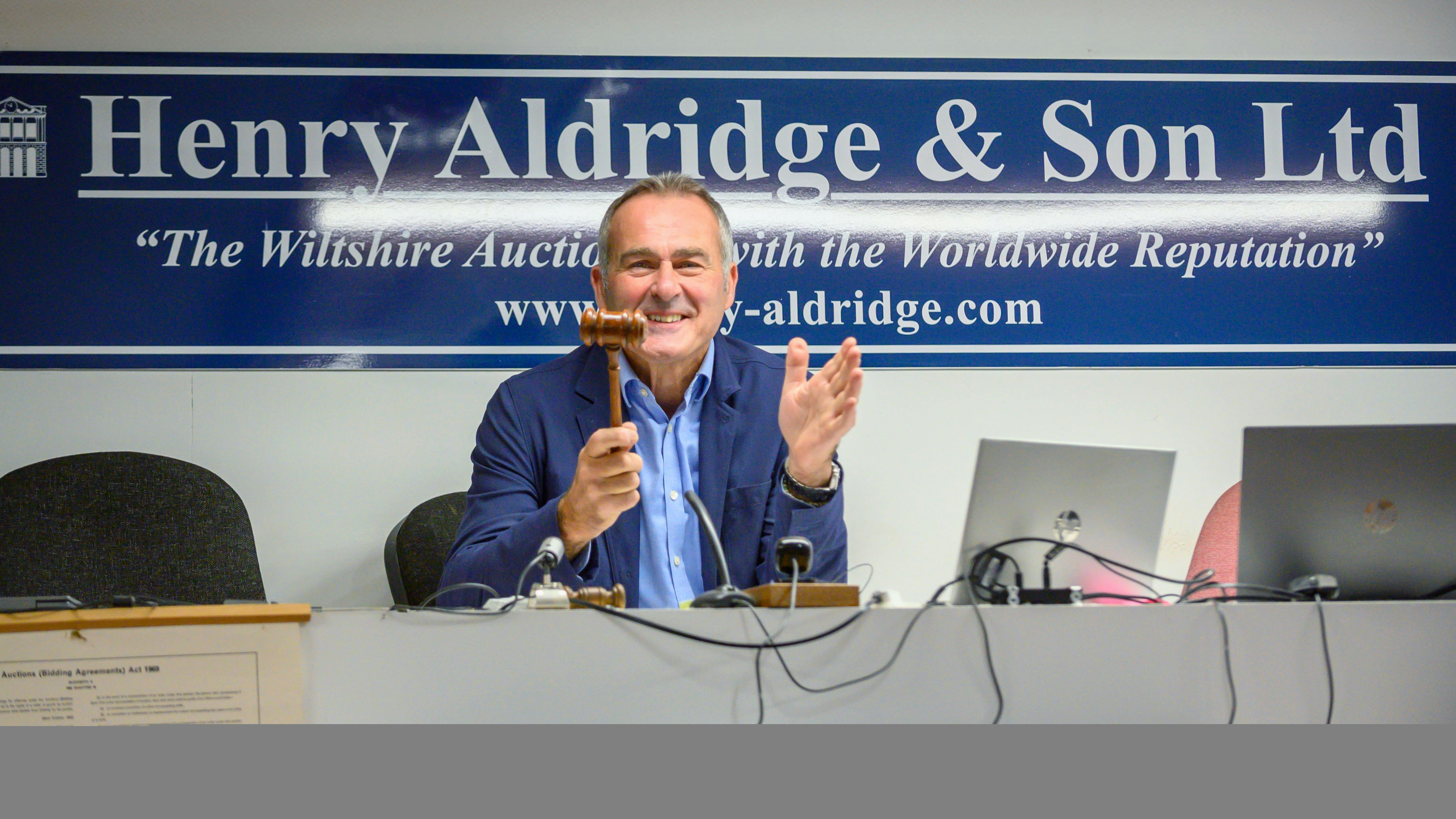 Auctioneer smiling, holding a gavel at Henry Aldridge & Son Ltd, with laptops and microphones on the desk.
