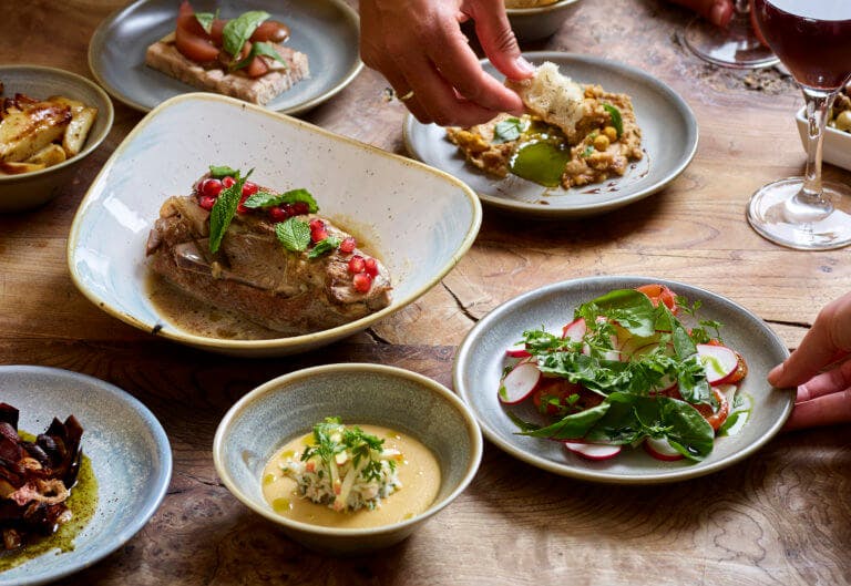 Selection of fresh seasonal dishes served on a wooden table with a hand dipping bread into chickpea dip