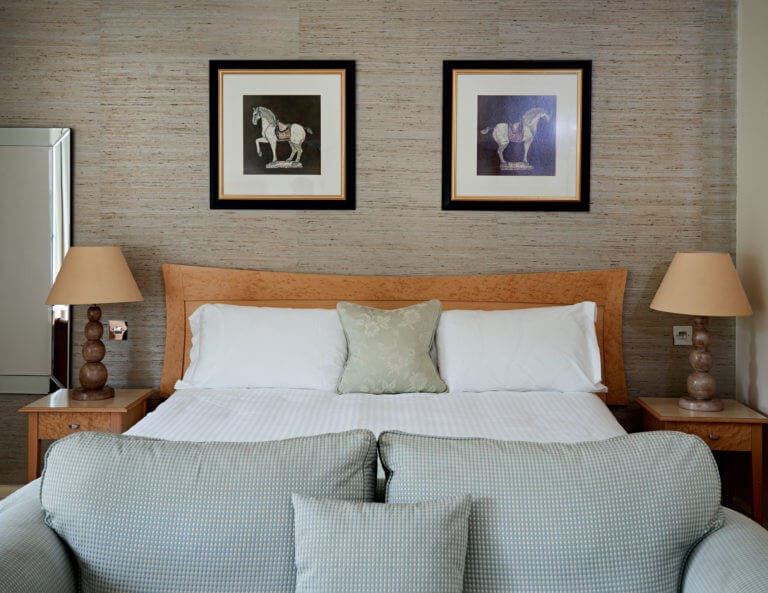 Hotel bedroom view of bed with wooden headboard, small gingham patterned sofa, bedside tables with large lamps and horse artwork above bed
