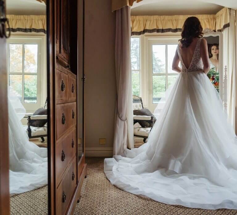 A bride in a a bedroom filled with period features admiring her wedding gown in a full length mirror.