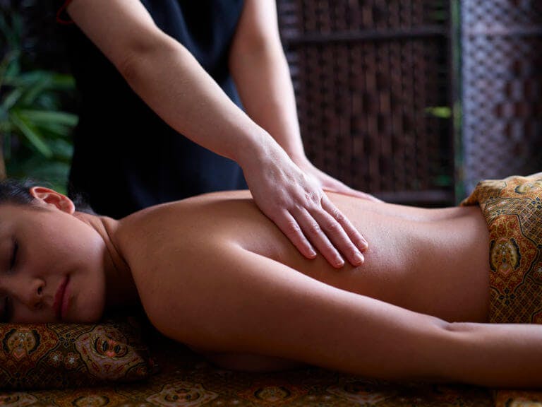 a female lying on her front under Thai patterned sheet receiving a back massage.