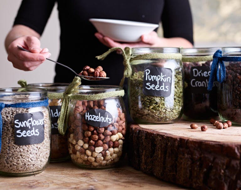 Jars of seeds and nuts in breakfast buffet on display, with person scooping some hazelnuts into a bowl