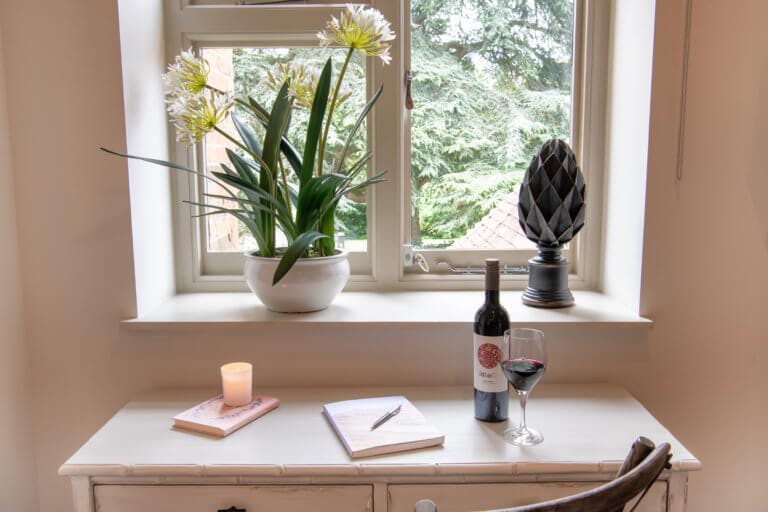 Desk with a bottle of red wine and filled wine glass, notepad and pen, lit candle on a book and window with flower plant on window sill