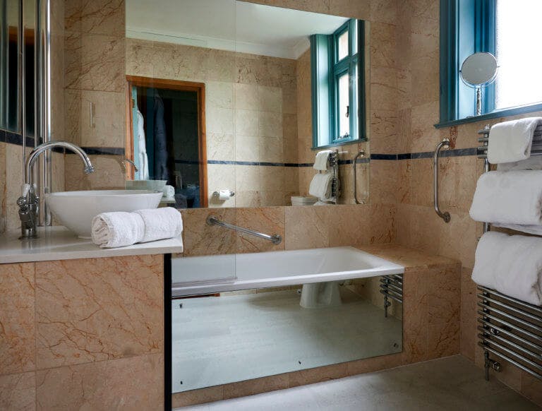 Bathroom in hotel room with mirrored bath edge, sink, large mirror above bath and marble effect tiles