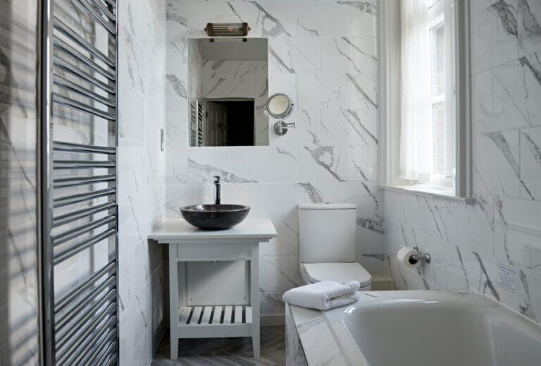 Small bathroom in Careys Manor Hotel manor suite with bath tub, toilet, sink and mirror above sink. Marble effect walls