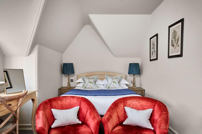 Small double hotel room with red armchairs in the foreground and bed in background with blue lamps either side