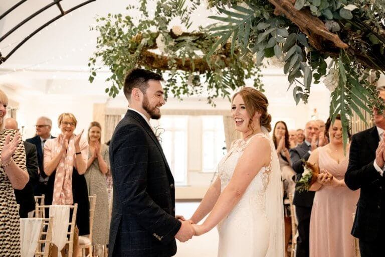 The bride and groom smile during the ceremony with guests clapping in the background