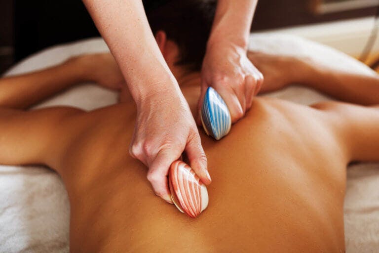 Man lies face down on massage bed as therapist runs two shells down the middle of his back