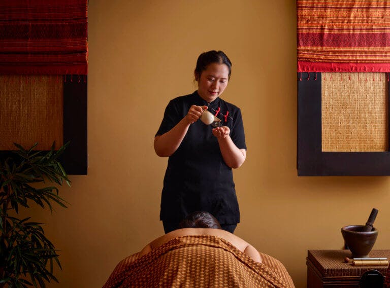 A spa therapist pouring oil from a small white jug into the palm of their hand. A female is lying on her front on a massage couch under Thai patterned fabrics.