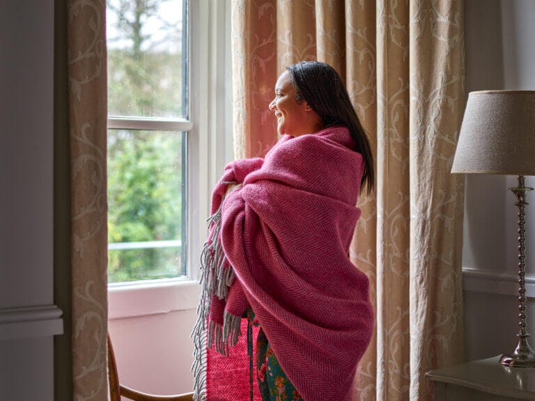Woman stares out of bedroom window at trees from Careys Manor Hotel room, wrapped in cosy pink blanket and wearing green silk Their Nibs pyjamas