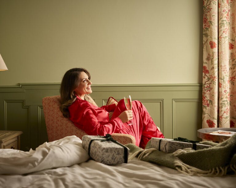 Woman in silk red festive pyjamas sits in an armchair next to a bed with Christmas gifts on it at Careys Manor Hotel in the New Forest