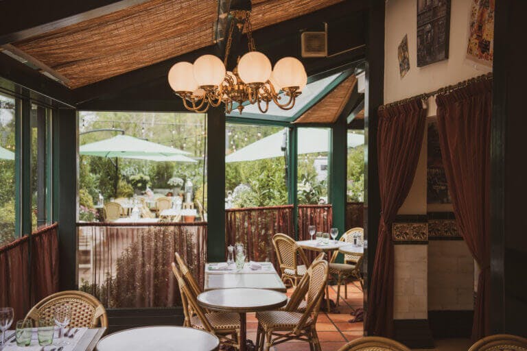 Inside Le Blaireau restaurant in Brockenhurst with small tables and rattan chairs, burgundy chiffon over windows looking out to al fresco dining area