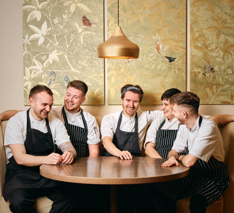 Group shot of happy chefs at Careys Manor hotel sitting at table in Cambium restaurant in their work uniforms with black aprons