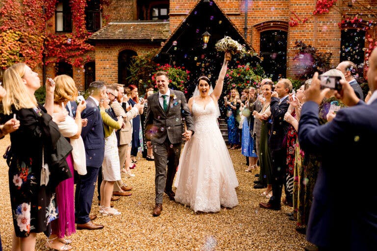 Bride and groom leave wedding ceremony surrounded by wedding guests blowing bubbles outside Careys Manor hotel