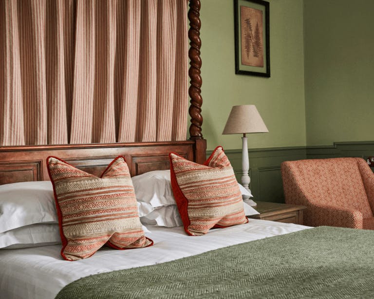 Close-up of an oak four-poster bed with striped drapes and cushions, a green wool throw and sage green walls.