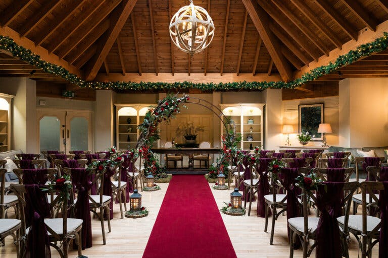 The lounge set for Maxine & Alastair's wedding ceremony at Hampshire wedding venue Careys Manor Hotel & SenSpa, photographed from the end of the aisle. A red carpet is placed down the aisle and a moon gate is behind the registrars table.