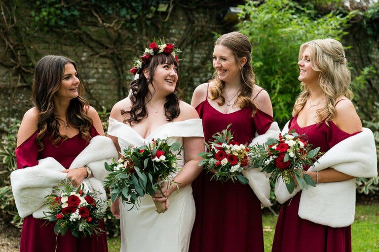 The bride smiles with her bridesmaids who wear red dresses with a fur shawl in the hotel gardens at Hampshire wedding venue Careys Manor Hotel & SenSpa.