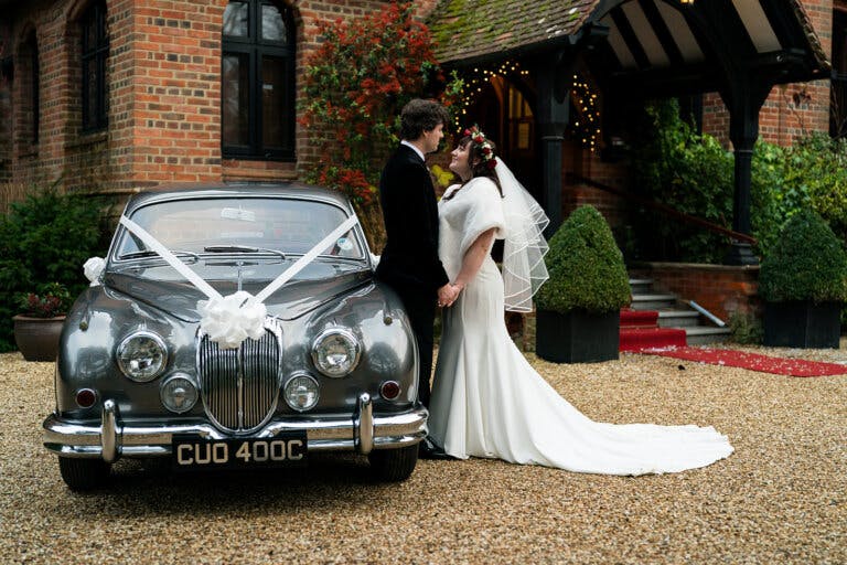 The bride and groom stand next to their vintage car at the hotels main entrance, kissing at Hampshire wedding venue Careys Manor Hotel & SenSpa.