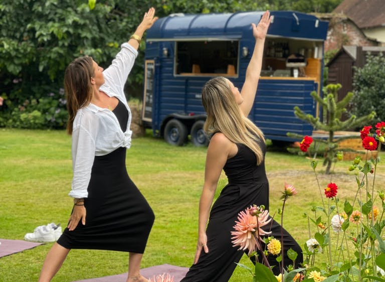 Two ladies on the front lawn holding a yoga post with their arm to the sky and the Neigh Bar horse box in the background