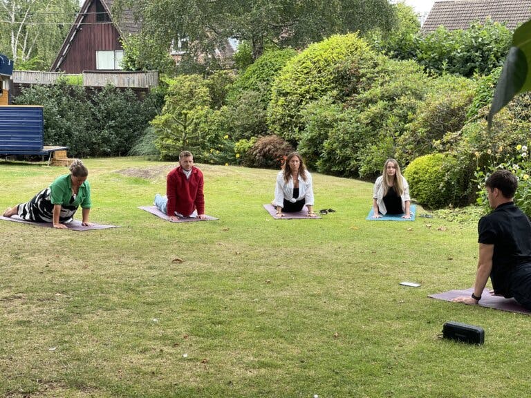 A group of people on the front lawn holding a yoga pose facing the instructor.