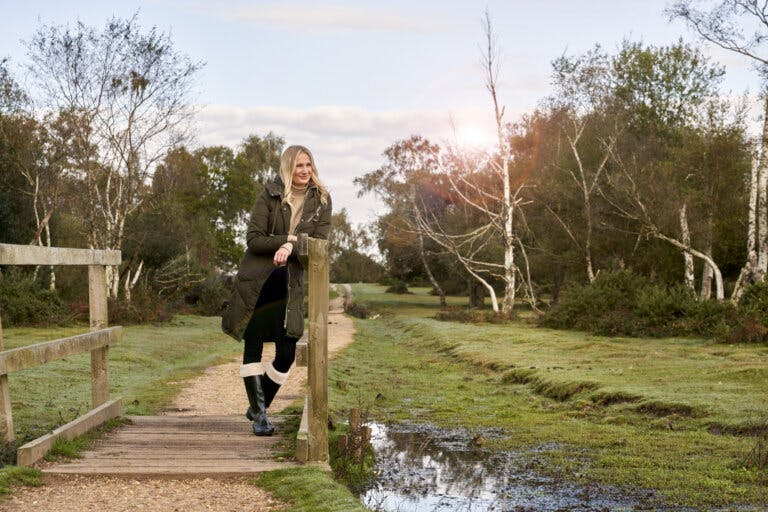 A blonde woman dressed in winter gear with wellies, jumper and coat stands on a wooden bridge in the New Forest national park looking out at the view