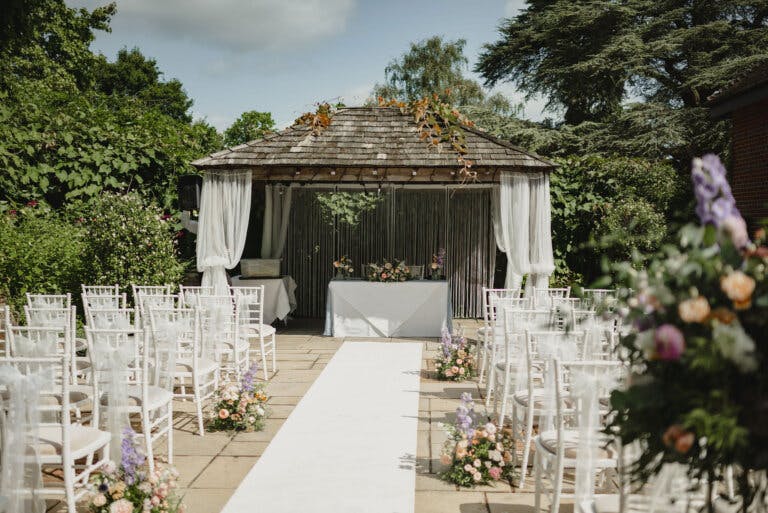 View down the white carpet aisle to the wedding wooden gazebo draped in white with chairs