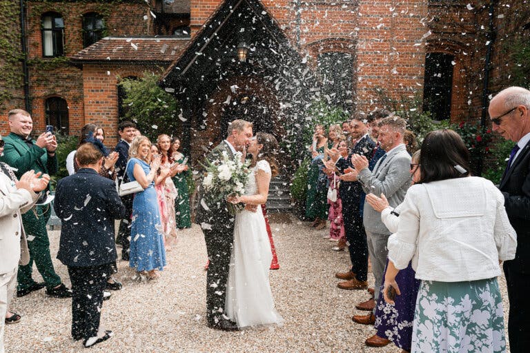 The newly married couple stand outside the main entrance with their guests either side, throwing confetti. The couple are kissing