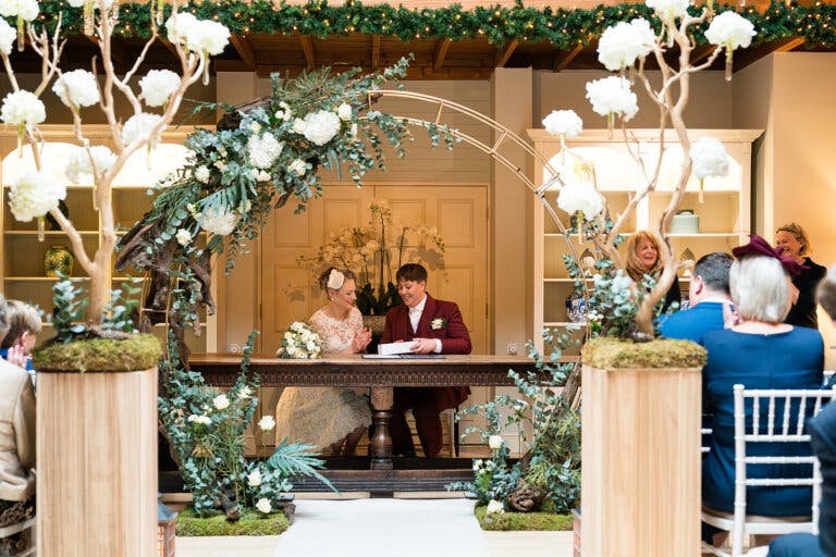 The brides sit at the registrars table with the floral moon arch around them