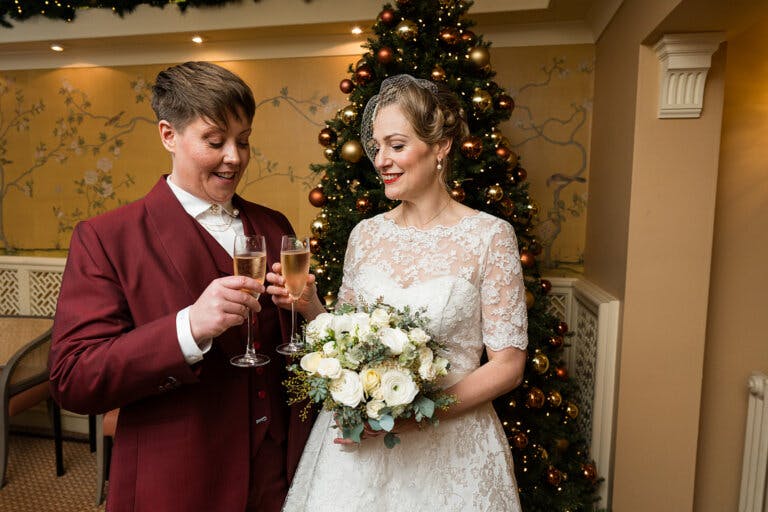 The Brides stand in front of a Christmas tree after the ceremony, clinking their champagne flutes.