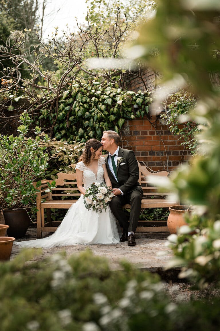 The bride and groom sit on a bench in the garden, the groom kisses the brides forehead