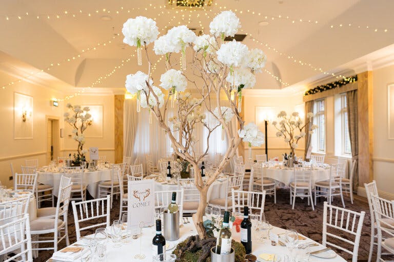 Image shows the Cedar Suite dressed for the wedding breakfast with tall white floral arrangements which are bud vases on a tree like structure