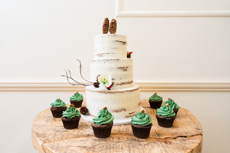 Image shows a 3 tier white wedding cake with rustic decoration and green cupcakes in front