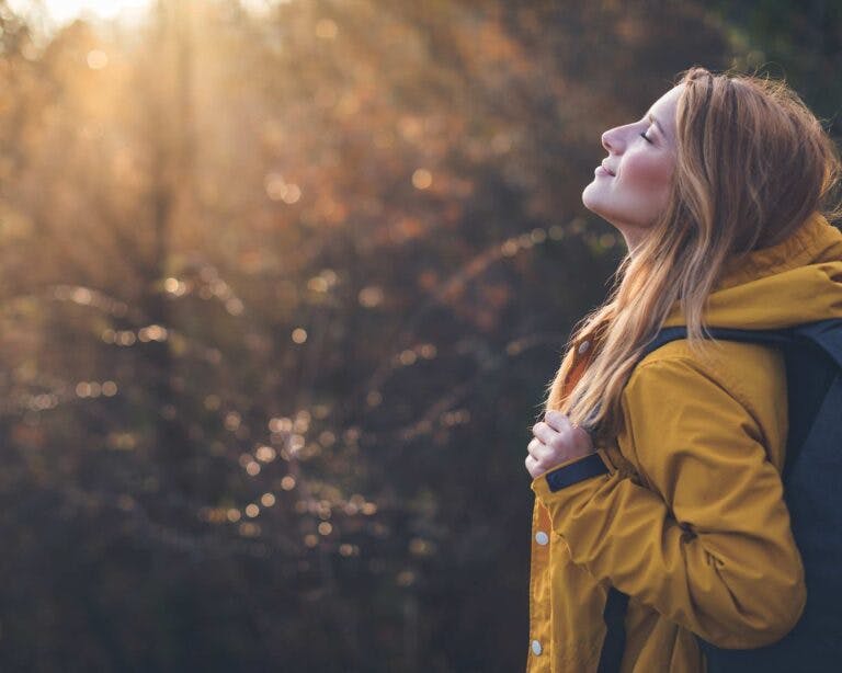 Woman stands in autumnal forest eyes closed breathing in fresh air wearing yellow raincoat and backpack