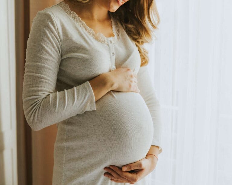Pregnant woman stands beside a white curtain in grey nightie cradling belly
