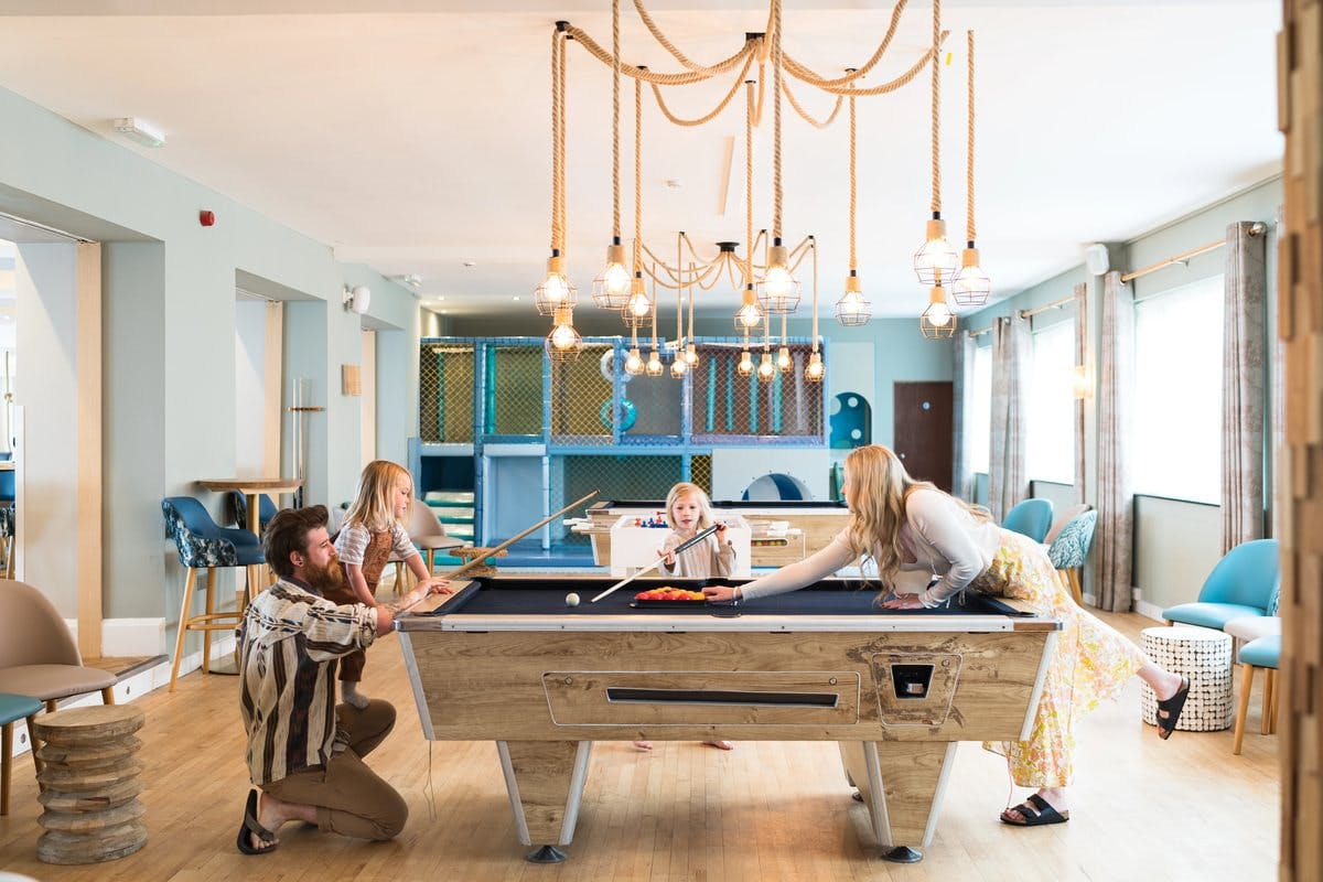 Family playing pool in a bright room with modern decor and hanging lights.