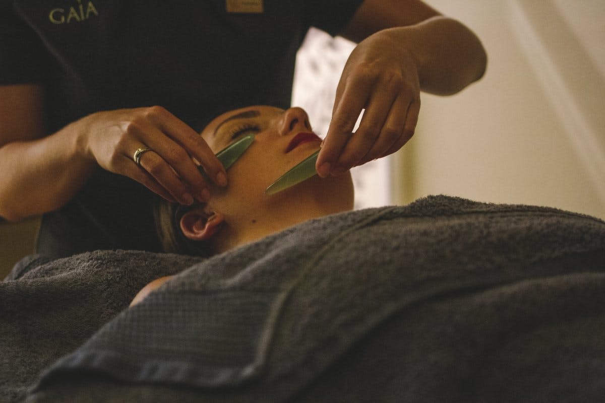 Person receiving a facial massage with jade rollers, lying under a towel.
