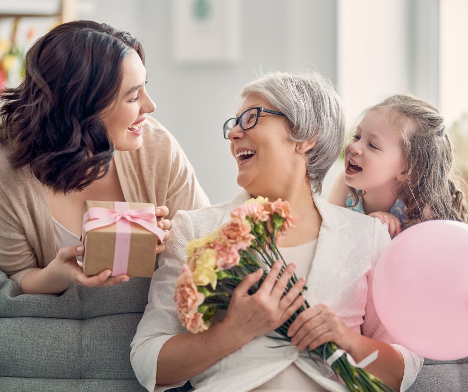 A joyful older woman with glasses holds flowers, sitting between a smiling young woman giving a gift and a laughing child with a pink balloon. Mother's Day
