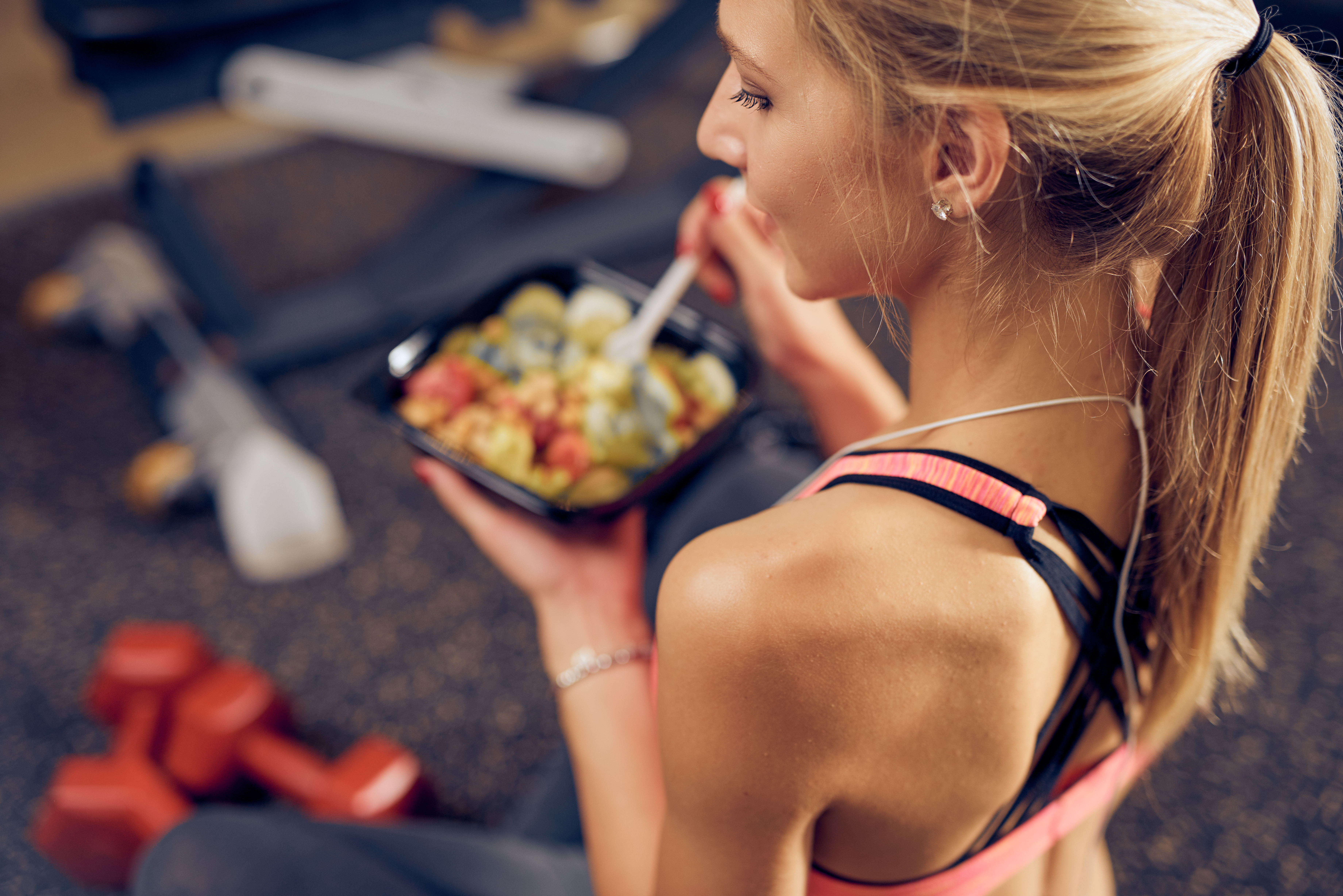 Young woman with tied back blonde hair sits in the gym, eating a bowl of high-carbohydrate salad as one of the best pre and post workout foods.