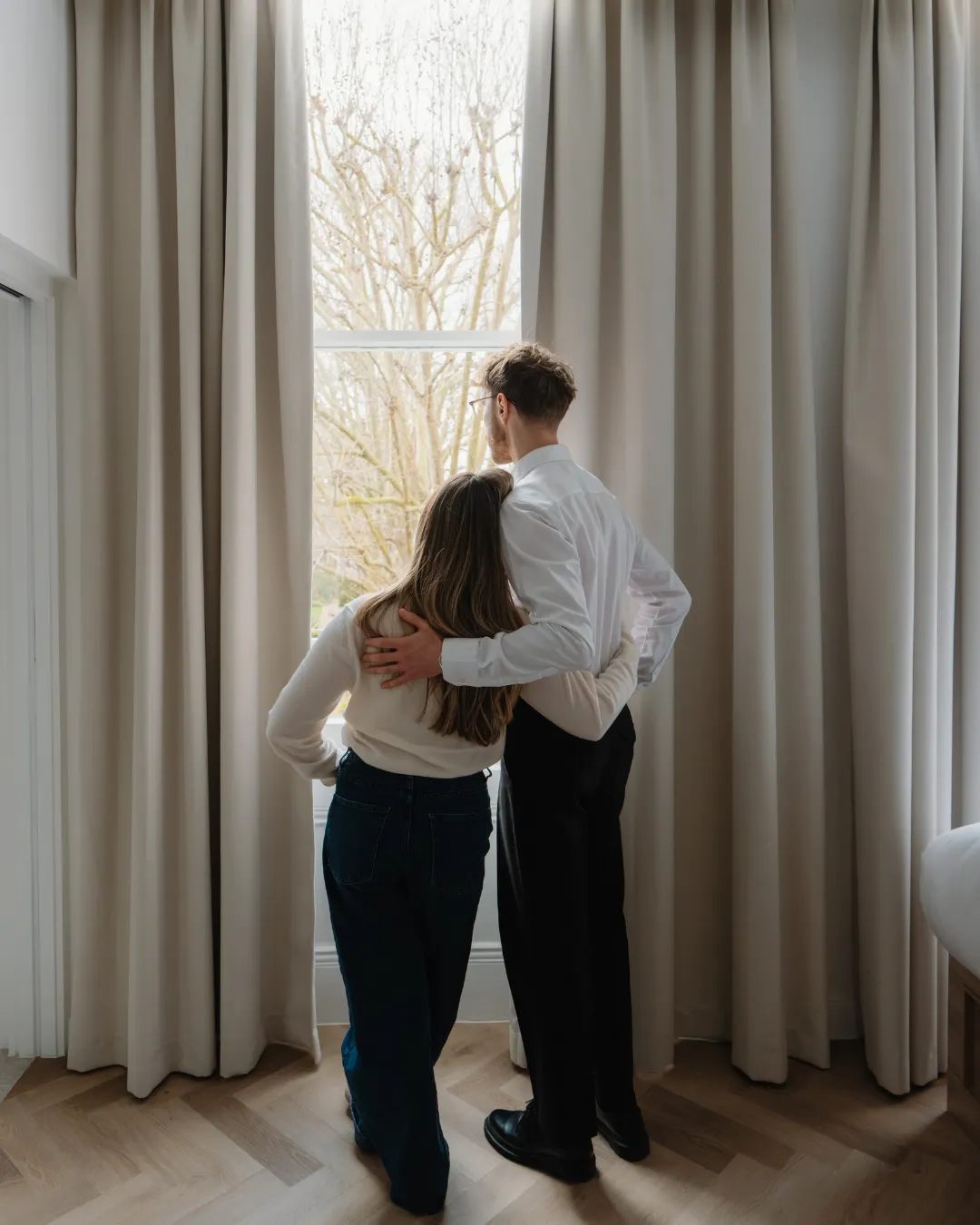 Two guests stand with their arms around each other looking out a tall window in a bright hotel room.