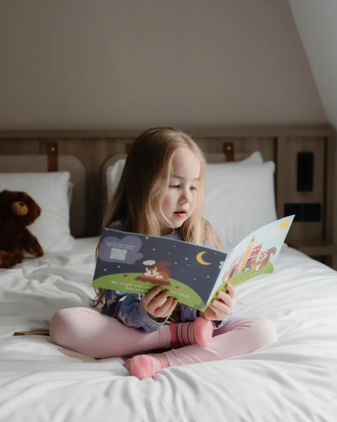 A young girl sits cross legged on a white hotel bed reading a book with a teddy bear behind her.