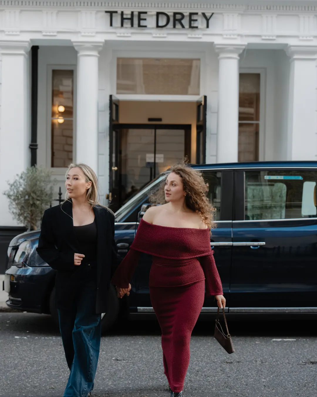 Two women hold hands outside The Drey with a black taxi parked behind them. Visible text reads "THE DREY".