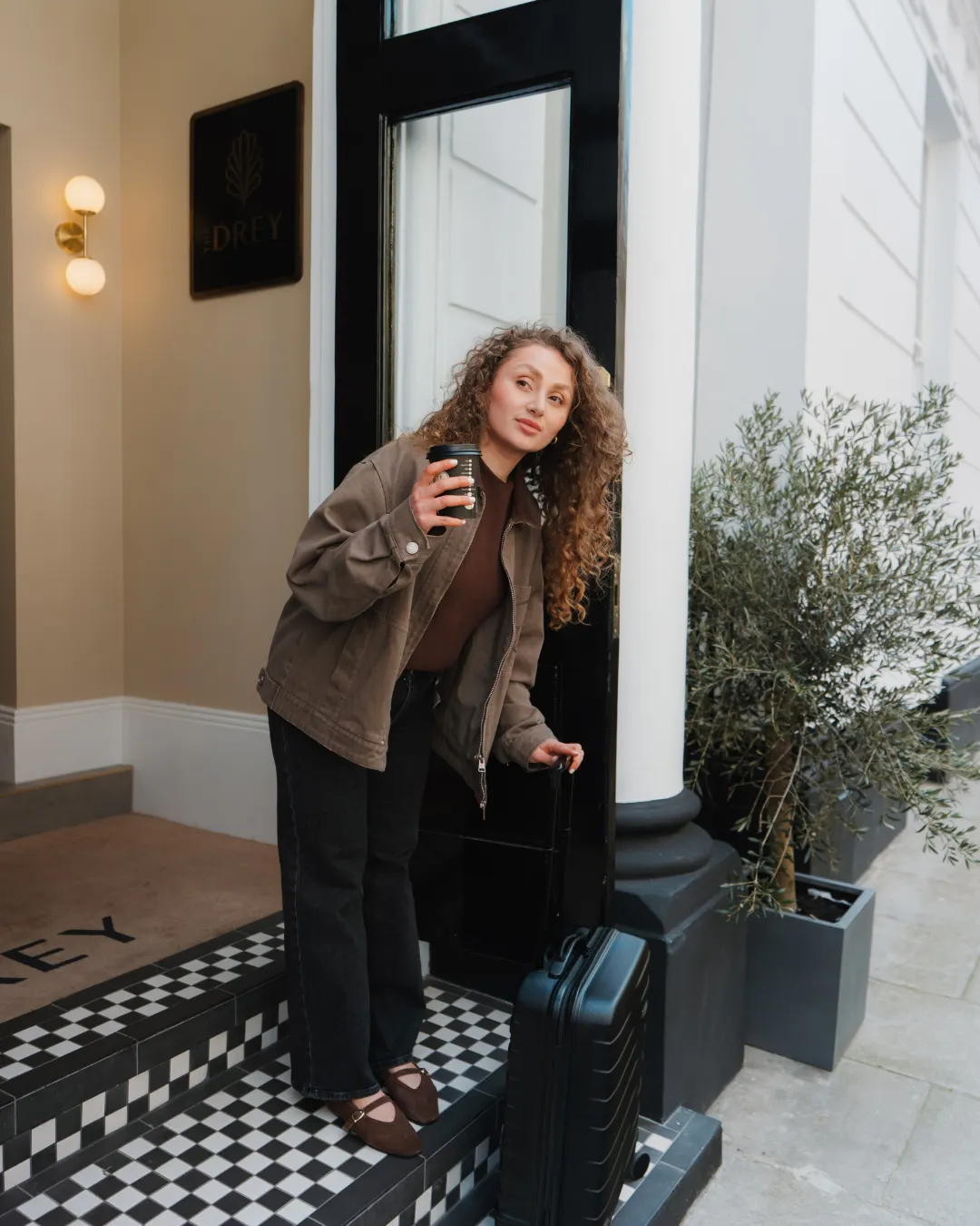 A guest stands on the checkerboard entrance steps at The Drey holding a coffee and a suitcase. Visible text reads "DREY".