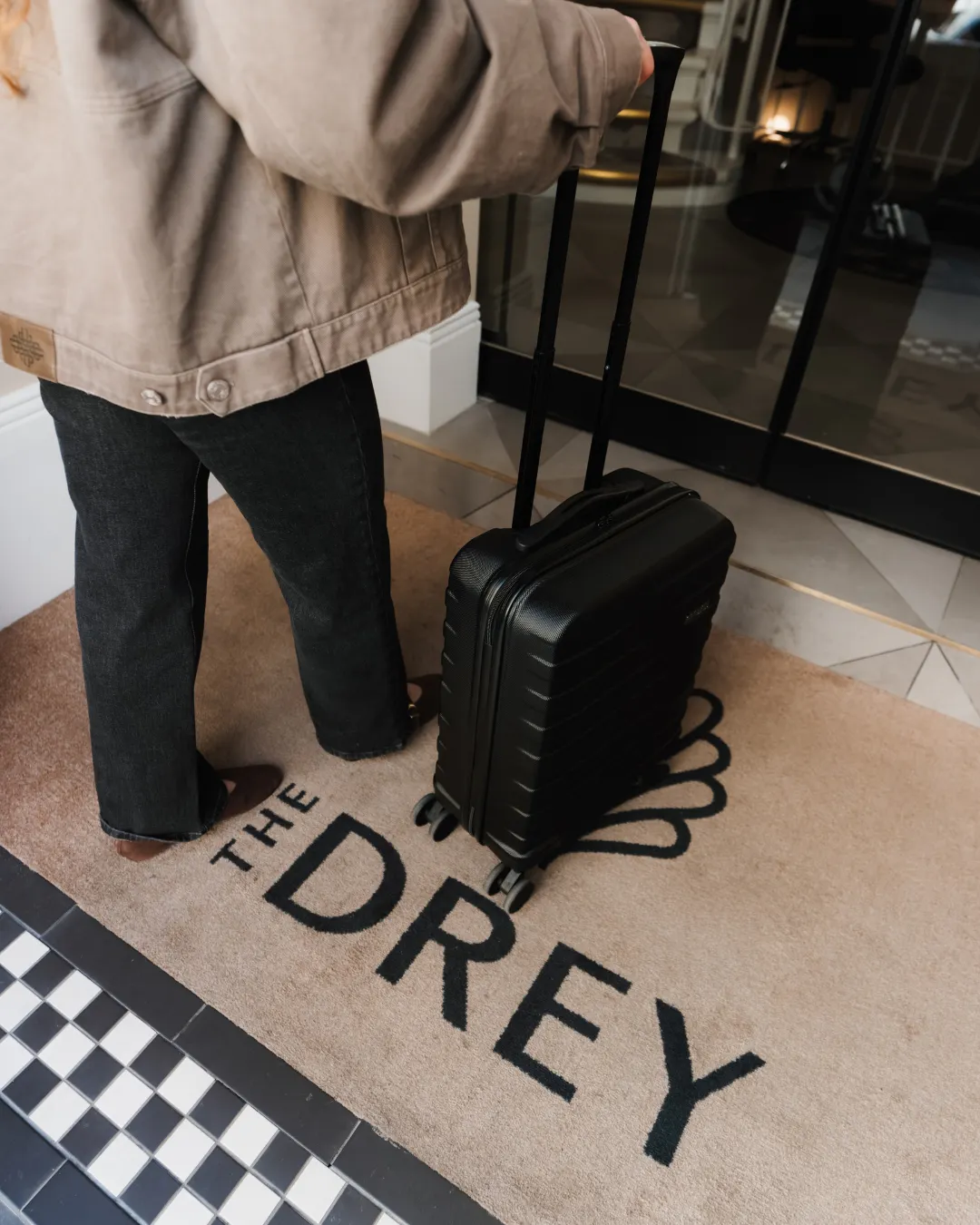 A guest stands on The Drey entrance mat with a black suitcase by the open doors. Visible text reads "THE DREY".